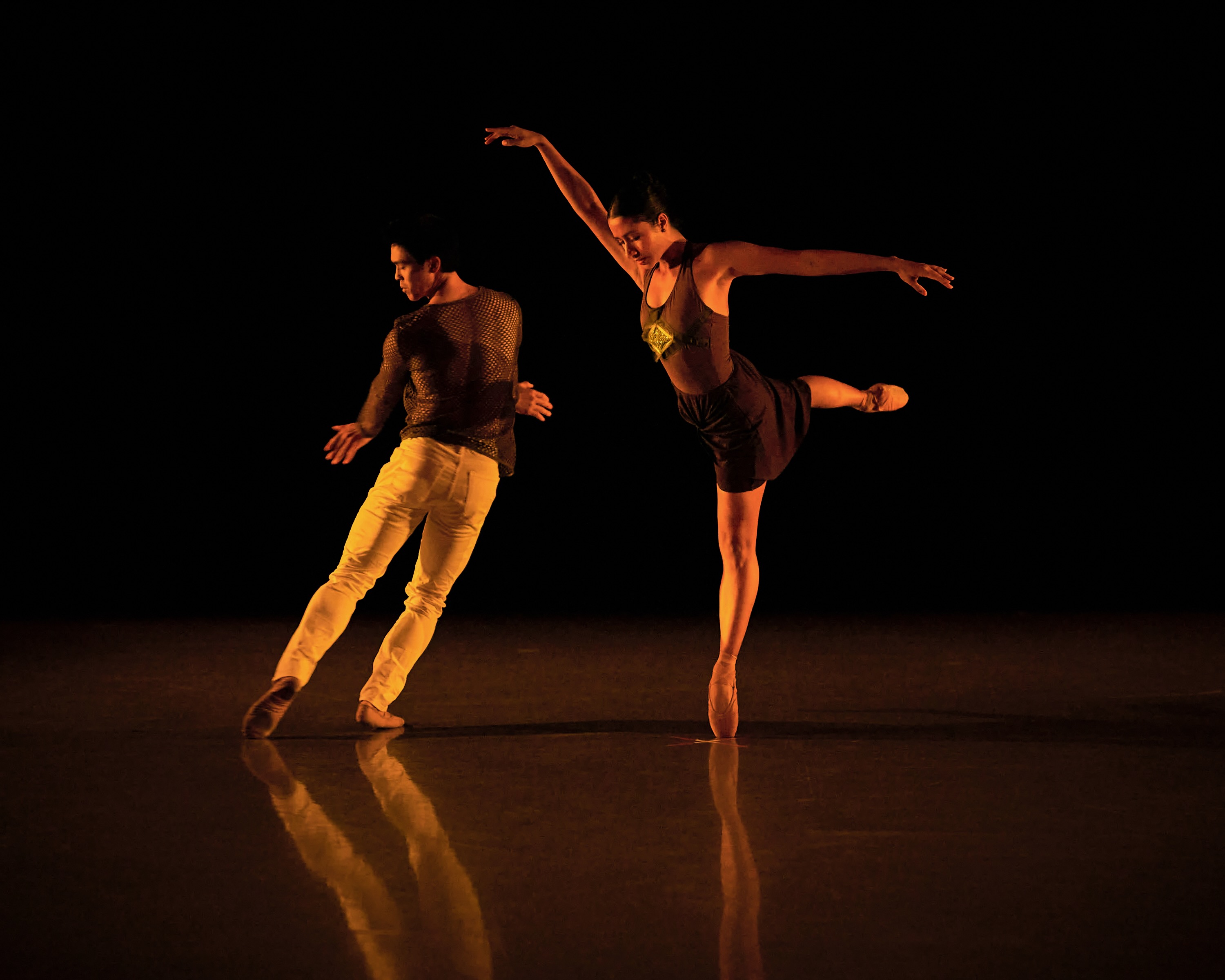 Two dancers move side by side in a pool of warm stage light.