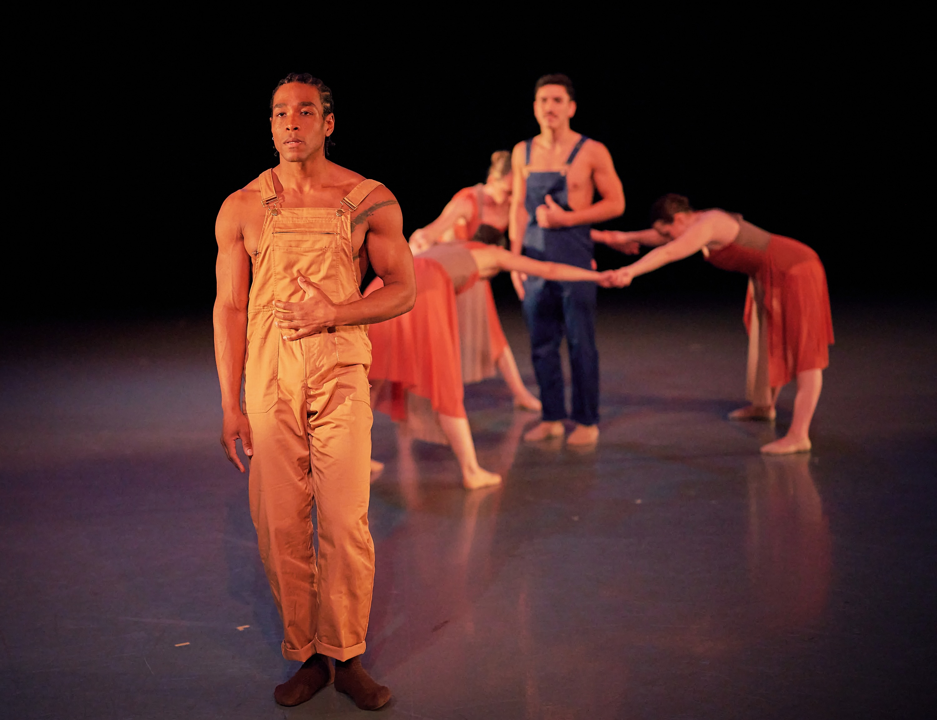 A dancer in tan overalls stands in the foreground as an ensemble unfolds behind him.