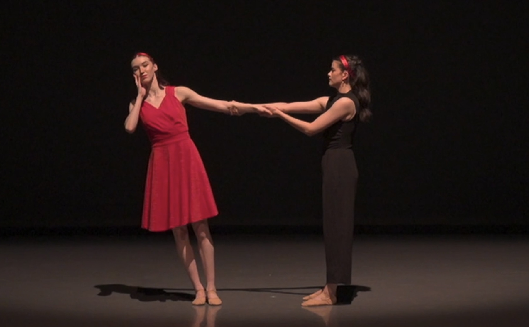 Two dancers in red and black costumes are connected through their hands on a dark stage.
