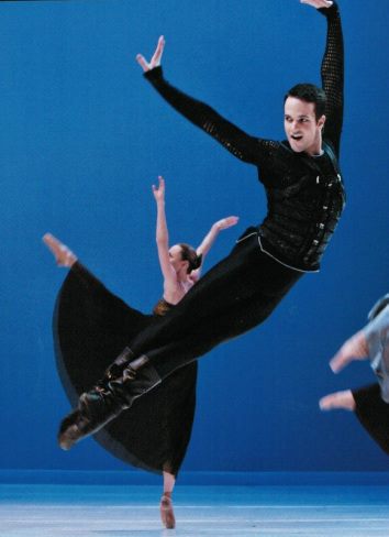 Ian Parsons airborne in a ballet performance against a blue-lit stage, with one arm extended overhead.
